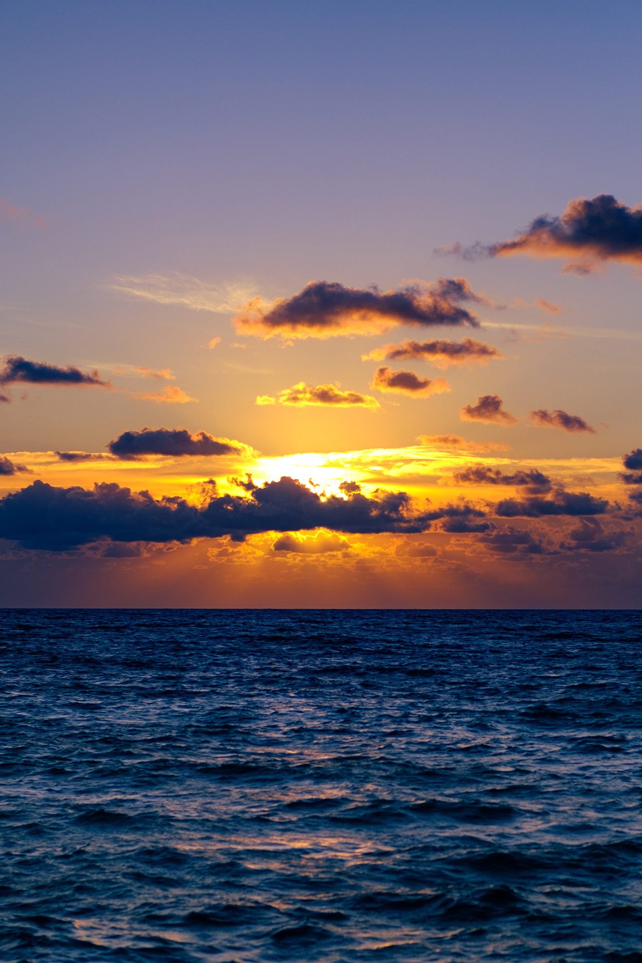 Sunset over the ocean with a vibrant sky. The sun is partially hidden behind clouds, creating a dramatic effect of orange and yellow hues reflected on the water below.