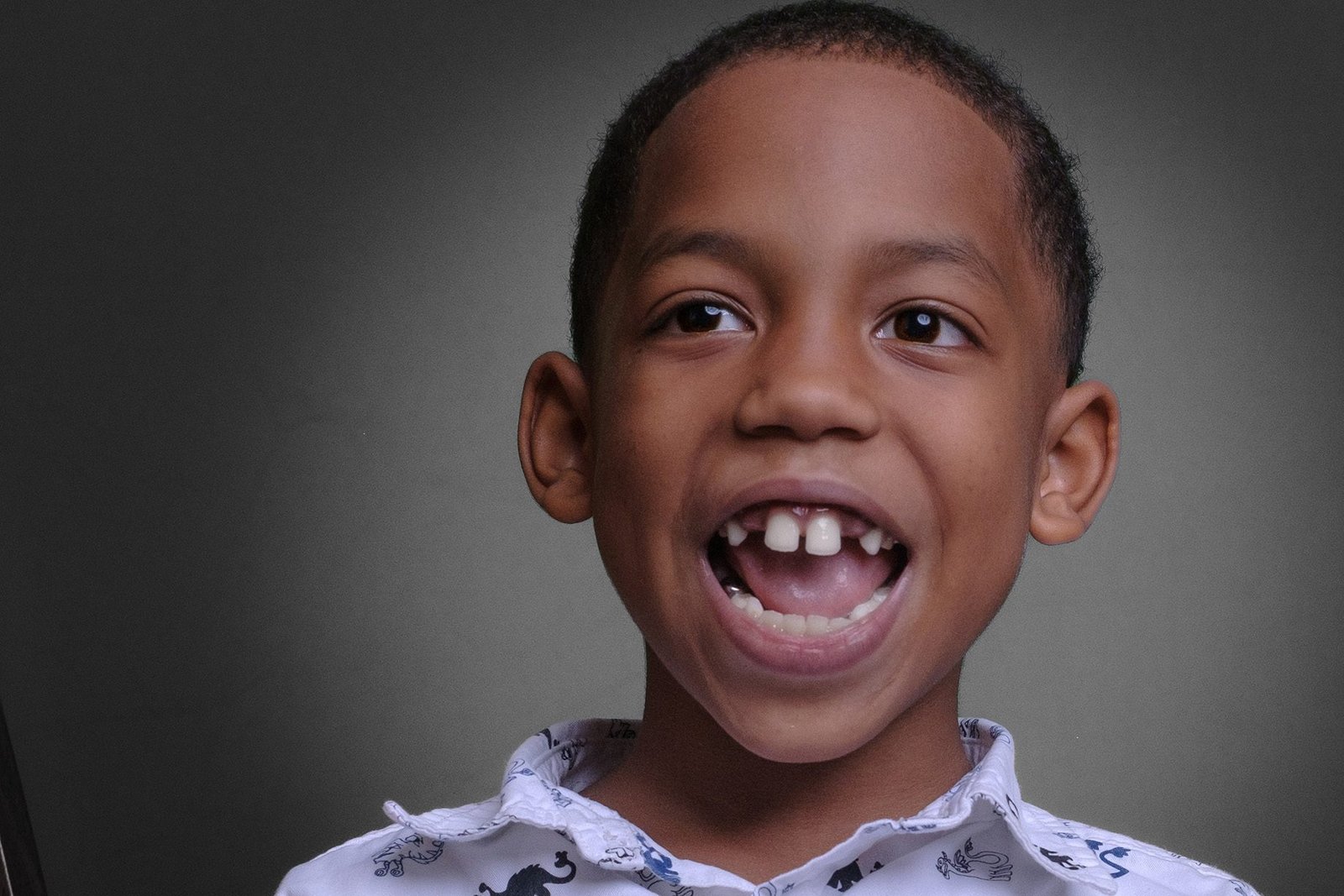 A young boy with a big smile and missing front tooth, wearing a white shirt with animal prints, against a grey background.