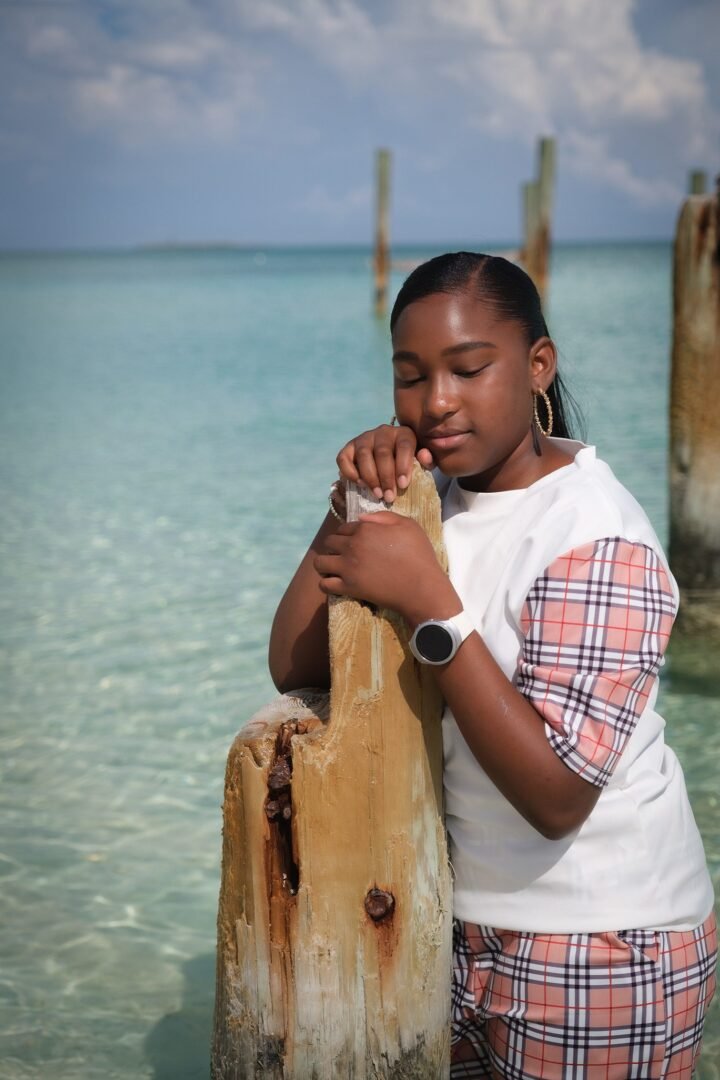 A young person stands by a wooden post in clear, shallow water, with eyes closed and a serene expression. Theyre wearing a white shirt with plaid sleeves and matching shorts, accessorized with hoop earrings and a smartwatch. The sky is partly cloudy.